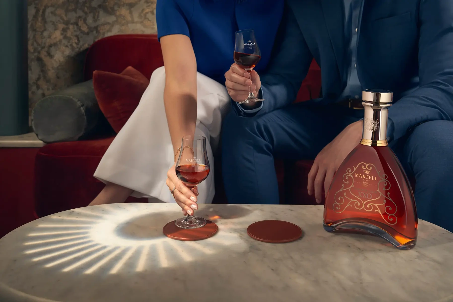 A man and woman seated on a velvet sofa enjoy glasses of Martell XXO cognac, with warm light casting Le Cercle by Pernod Ricard's logo on a marble table beside the elegant cognac bottle.