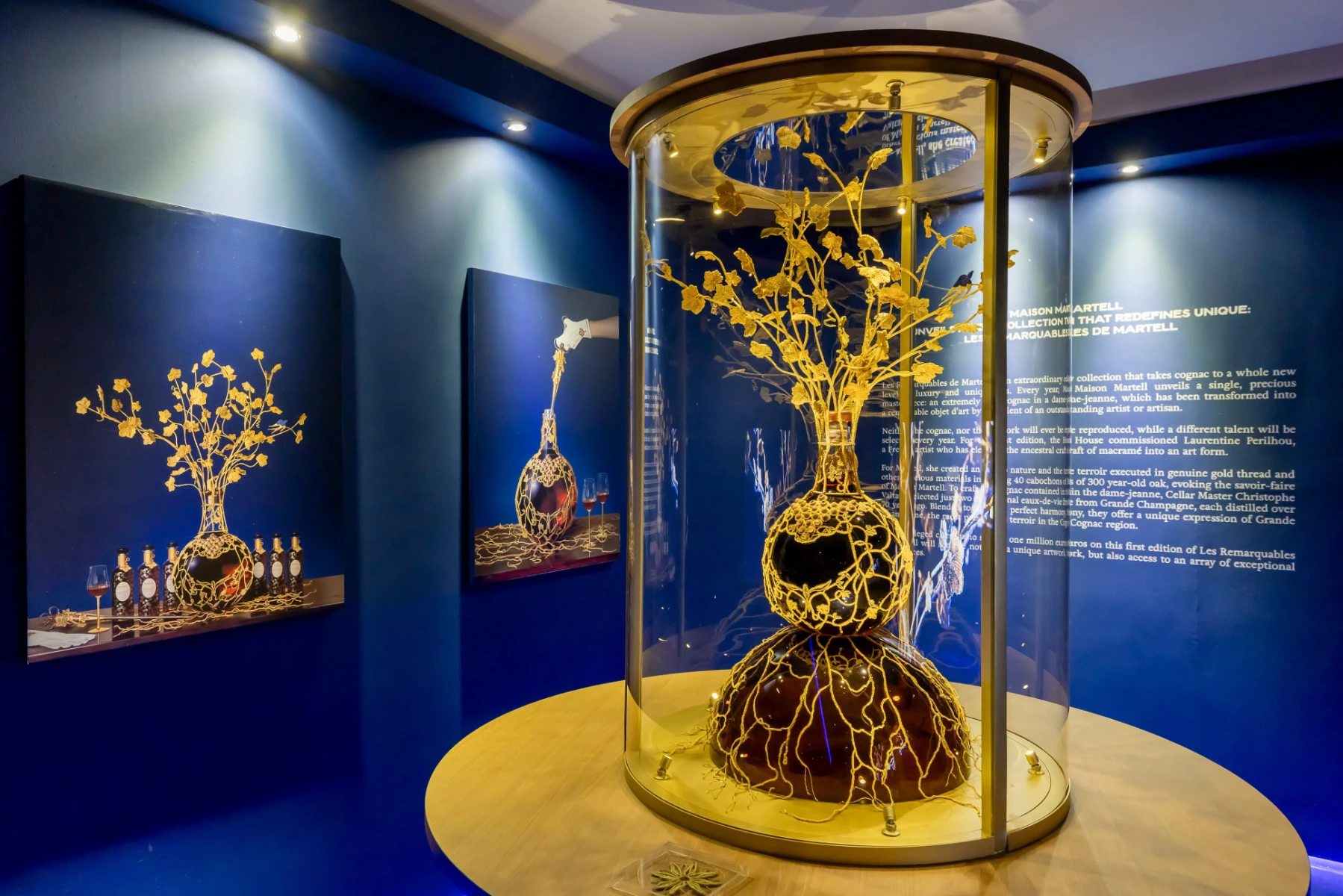 An illuminated exhibition of Maison Martell’s Les Remarquables collection, highlighting a handcrafted golden sculpture rising from a cognac decanter within a glass cylinder, presented against elegant blue walls with curated artwork and storytelling panels.
